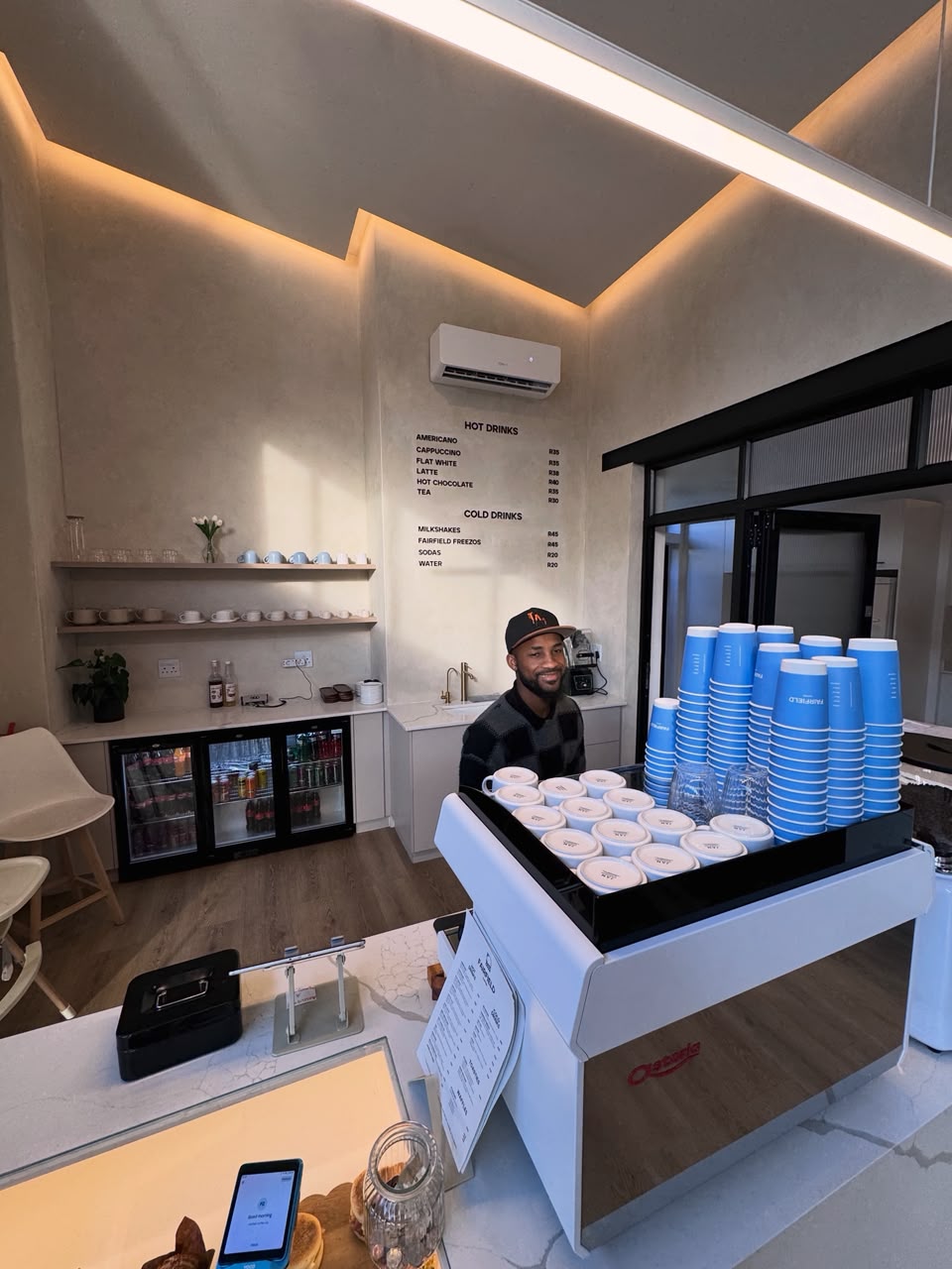 Barista behind the Fairfield Coffee counter with the hot and cold drinks menu on the wall